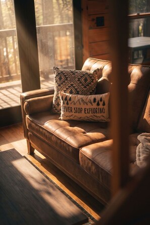 A leather sofa with a cushion that reads 'NEVER STOP EXPLORING' in a room with a wooden floor and a window. - Smokies Sunrise | Hot Tub, Fireplace & More! (Sevierville)