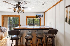 A kitchen with a bar area, wooden stools, a ceiling fan, and a view of the outdoors through large windows. - Smokies Sunrise | Cozy Rustic Cabin Experience (Sevierville)