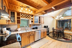 A kitchen with wooden cabinets, a black countertop, stainless steel appliances, and a dining area with a table and chairs. - Smokies Sunrise | Cozy Rustic Cabin Experience (Sevierville)