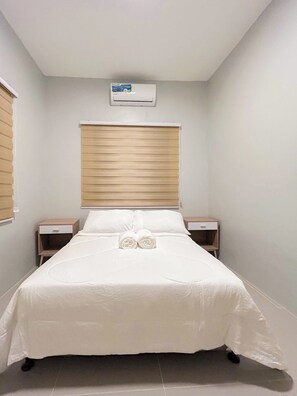 A neatly made bed with a white duvet and pillows, two bedside tables with lamps, a window with blinds, and an air conditioning unit on the wall.