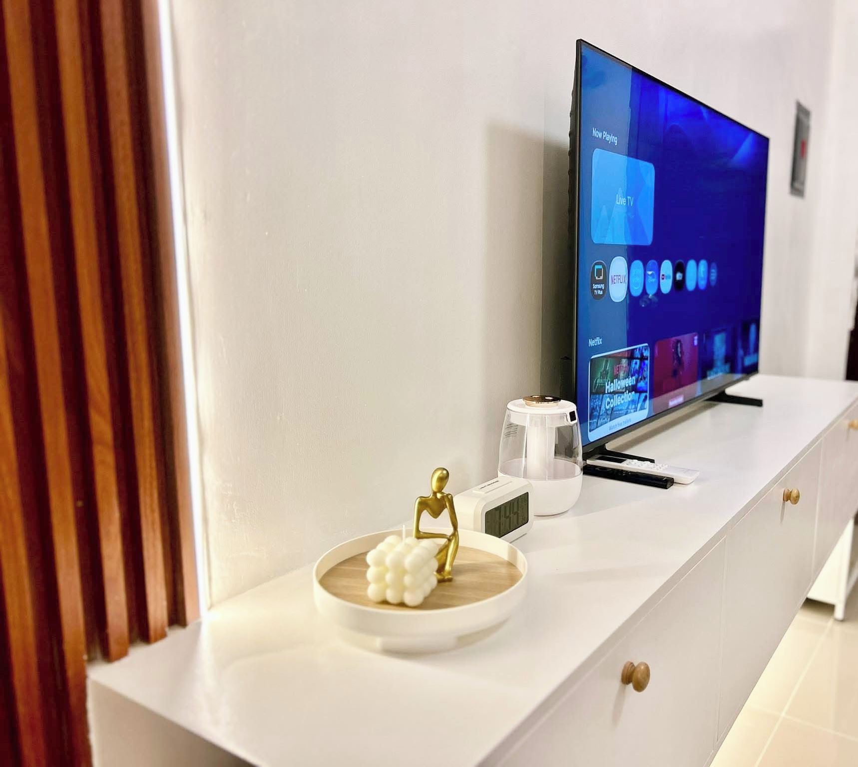 A modern living room with a flat-screen TV displaying a streaming service interface, a minimalist white console with a decorative figure and a clock, and a glass coffee table.