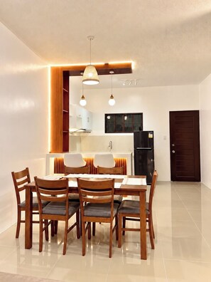 A dining area with a wooden table and chairs, a kitchen in the background, and a dark door.