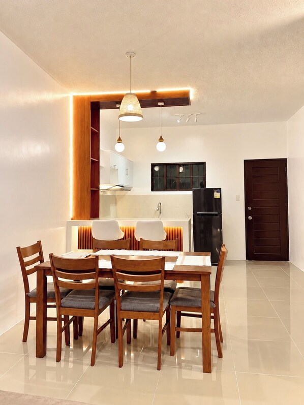A dining area with a wooden table and chairs, a kitchen in the background, and a dark door.