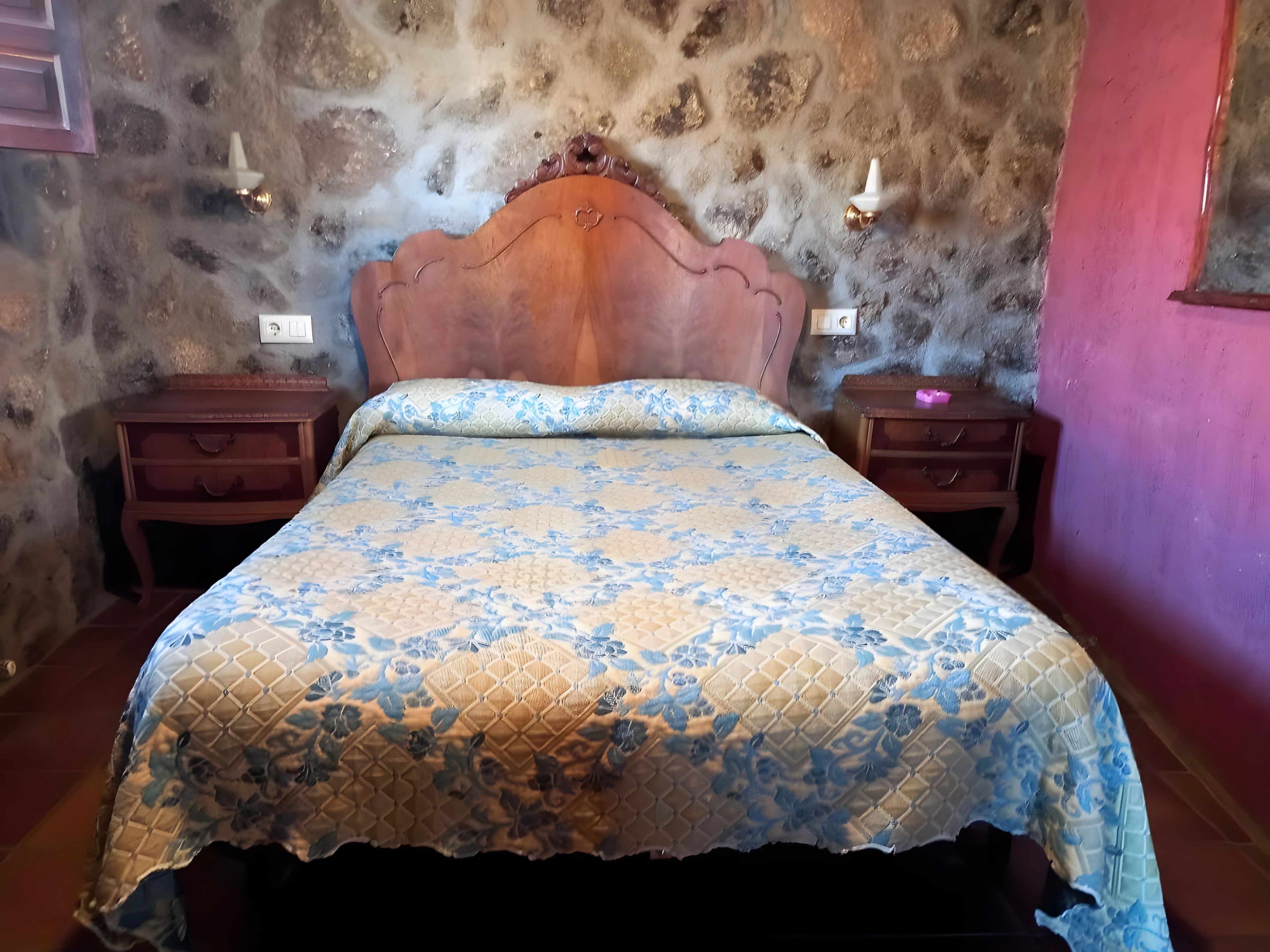 Ein Schlafzimmer mit einem hölzernen Kopfende, einer zudeckenden Bettdecke mit Blumenmuster, zwei Nachttischen und einer Steinwand.