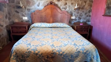 Ein Schlafzimmer mit einem hölzernen Kopfende, einer zudeckenden Bettdecke mit Blumenmuster, zwei Nachttischen und einer Steinwand.