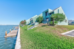 A waterfront property with a wooden dock extending into the water, a grassy area with a staircase leading to a building, and a clear blue sky. - First-Floor Waterfront Condo | Perfect Fishing Getaway and Great Views (Corpus Christi)