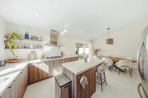 A modern kitchen with a central island, wooden cabinets, and a dining area with a table and chairs. - Modern 4BR Palm Beach Home | Garden & BBQ (Noord)