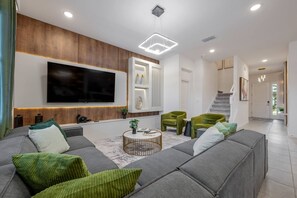 A modern living room with a large flat-screen TV, a grey sectional sofa, a round coffee table, and a staircase leading to another level. - 8BR Themed Villa • Pool, Game Room & Theater (Orlando)