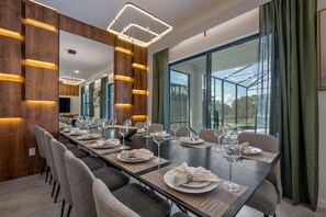 A modern dining area with a long table set for a meal, large windows, and a wooden accent wall. - 8BR Themed Villa • Pool, Game Room & Theater (Orlando)