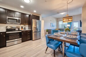 A modern kitchen with dark wood cabinets, stainless steel appliances, and a dining area with blue chairs and a wooden table set for a meal. - Vista Cay 403 • 3BR Condo, King Suite • Universal (Orlando)