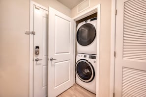 Une buanderie équipée d’un lave-linge et d’un sèche-linge à chargement frontal, d’une porte avec une poignée et d’une bouche d’aération située au-dessus du sèche-linge.