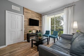 Een moderne woonkamer met een flatscreen televisie aan een houten wand, een zwarte salontafel, twee blauwe fauteuils, een grijze bank en een balkon met witte gordijnen.