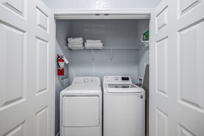 A laundry room with a front-loading washer and dryer, a fire extinguisher, and a shelf with folded towels. - Vista Cay 3BR Condo • King Suite • Near Universal (Orlando)