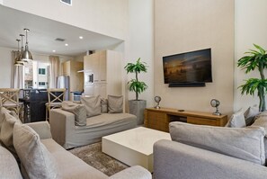 A modern living room with a sofa set, a coffee table, a TV mounted on the wall, and a dining area visible in the background. - 3 Suites • Private Hot Tub • Near Disney (Kissimmee)