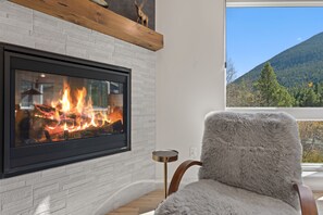 Ein moderner Kamin mit sichtbarem Feuer, ein bequemer Sessel und Blick auf einen bewaldeten Berg.