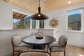 A dining area with a round table, four chairs, a hanging light fixture, and a view of a mountainous landscape through large windows. - Upscale 3BR Cambers Townhome w/ Private Balcony (Keystone)