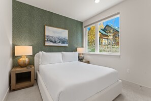 Ein Schlafzimmer mit einem Bett, zwei Nachttischen, einer Lampe, einem Gemälde und einem Fenster mit Blick auf ein Haus.