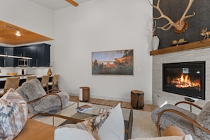 A modern living room with a fireplace, seating area, and a kitchen in the background. - Upscale 3BR Cambers Townhome w/ Private Balcony (Keystone)