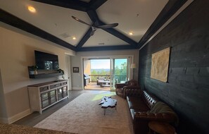 Un salón espacioso con una pared de acento de madera oscura, un sofá de cuero, un ventilador de techo y un gran ventanal con vistas al exterior.