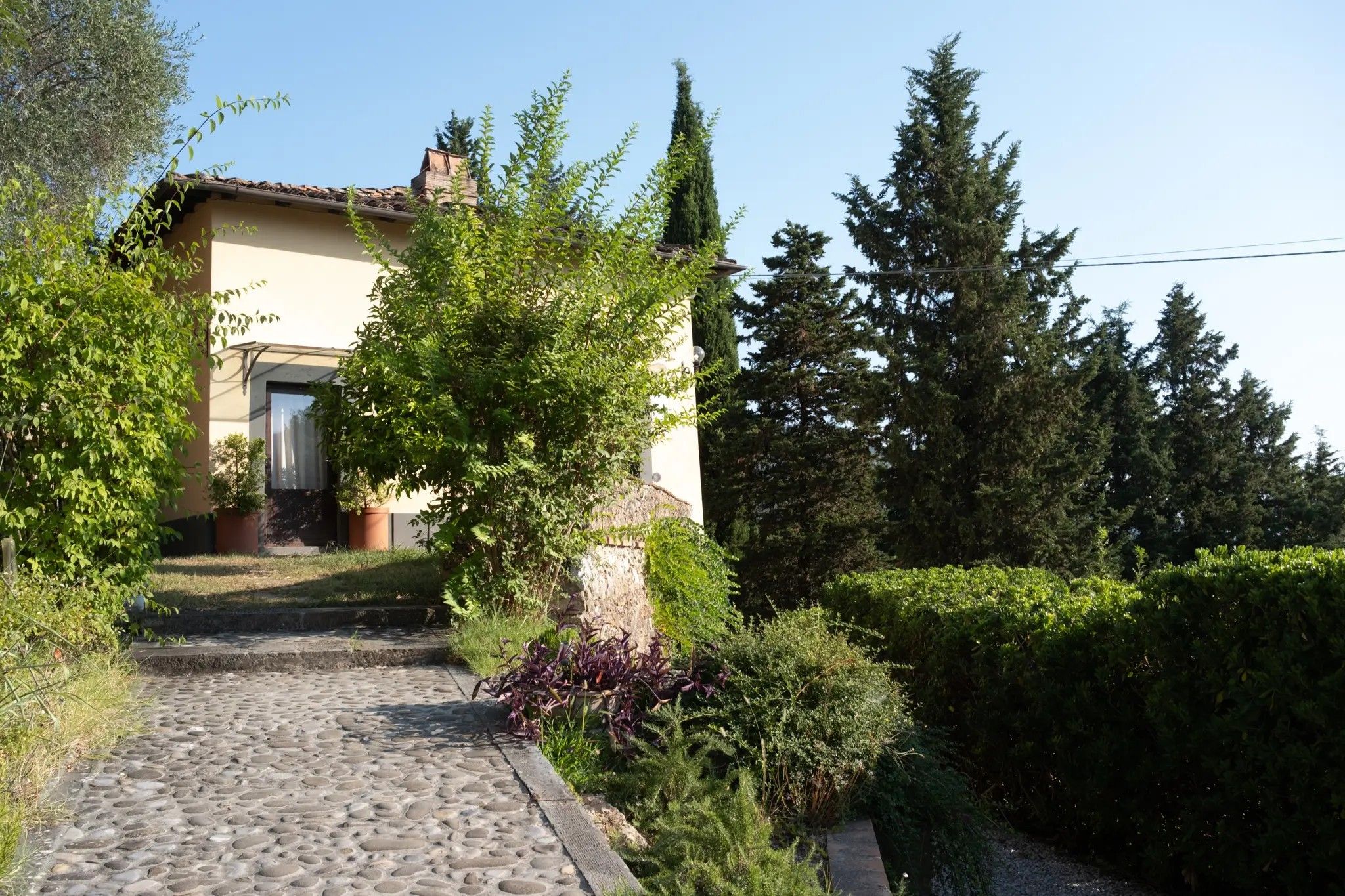 Un sentiero in pietra conduce a un edificio con tetto in tegole, circondato da vegetazione rigogliosa e alberi alti.