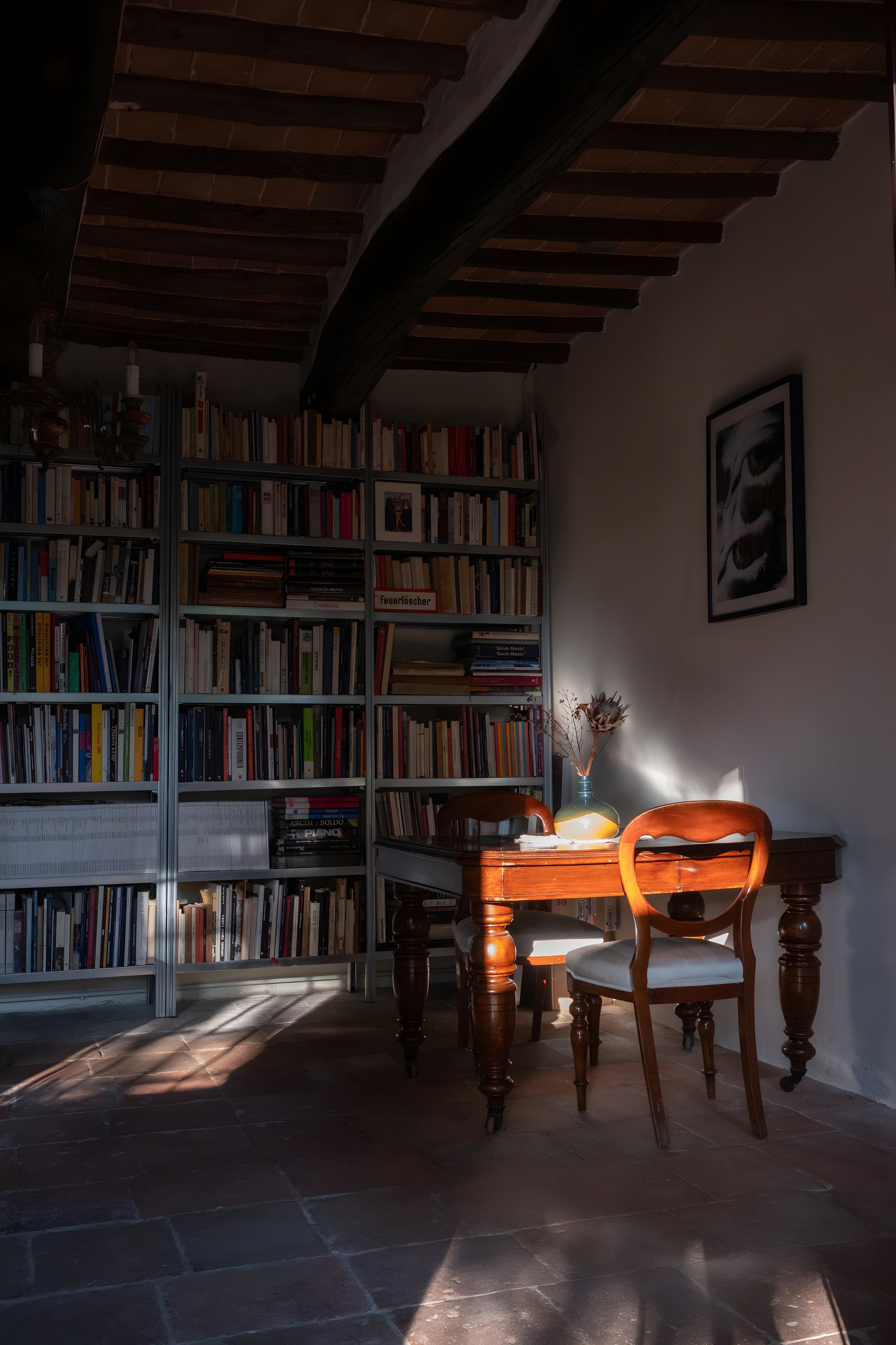 A room with a wooden ceiling, a bookshelf filled with books, a table with chairs, and a vase with dried flowers.