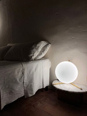 A bed with a white headboard and pillows, a bedside table with a lamp, and a wall with a textured surface. - Room 'The Mediterranean' with Mountain View, Wi-Fi and Air Conditioning (Lucca)
