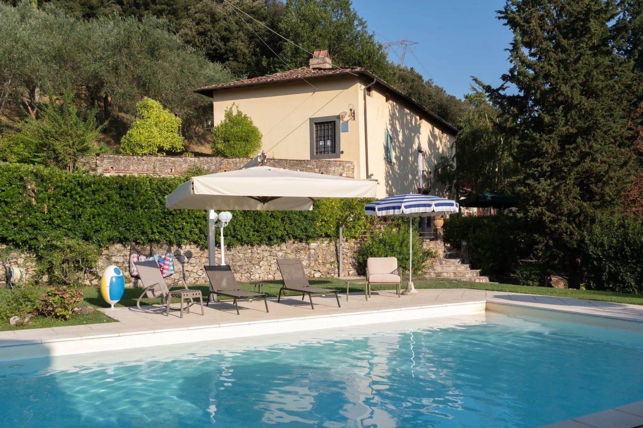 A swimming pool with lounge chairs and umbrellas, surrounded by a stone wall and greenery.