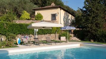 A swimming pool with lounge chairs and umbrellas, surrounded by a stone wall and greenery.