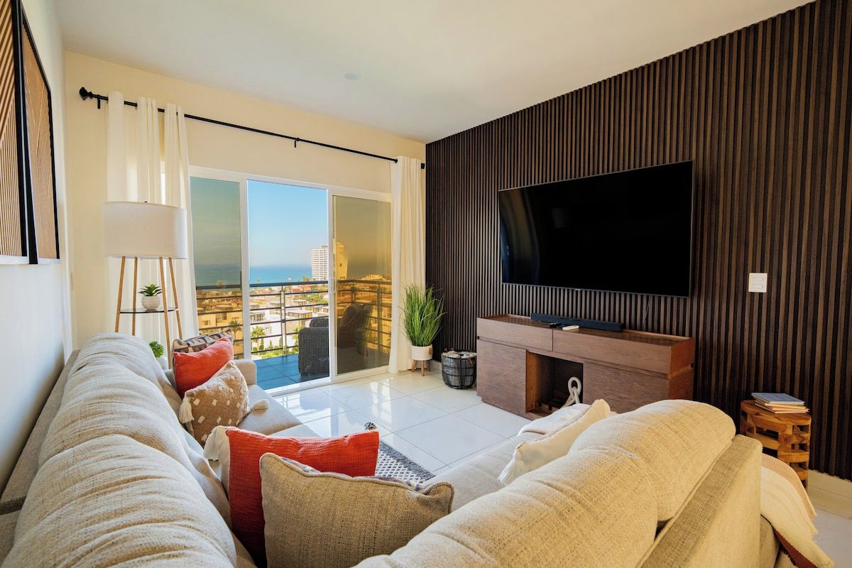 Sala de estar moderna com sofá, televisão de tela plana e porta de vidro deslizante que dá para uma varanda com vista da cidade.