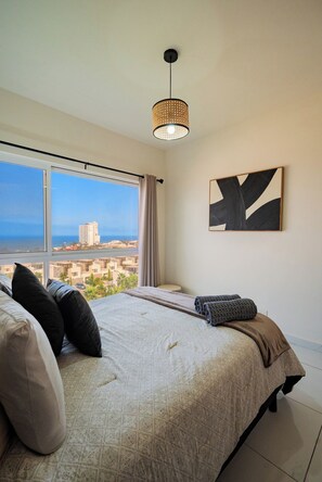 A bedroom with a large bed, a view of the city, and a modern pendant light. - Apartament 3Bd Amaralta (Playas de Rosarito)