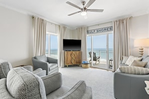 A living room with a ceiling fan, a flat-screen TV, and a view of the ocean through large windows. - Beachfront Corner Condo | 3BR/3BA | Beach Gear | 2 Parking Passes (Gulf Shores)
