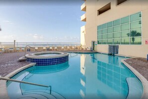 A swimming pool with a hot tub, surrounded by a brick patio and a building with large windows. - Beachfront Corner Condo | 3BR/3BA | Beach Gear | 2 Parking Passes (Gulf Shores)