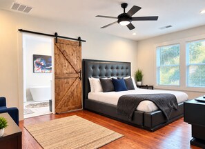 A bedroom with a large bed, a ceiling fan, a sliding barn door, and a view of the outside through large windows.