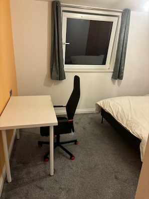 Una habitación pequeña con un escritorio blanco, una silla de oficina negra, una cama con edredón blanco y una ventana con cortinas grises.