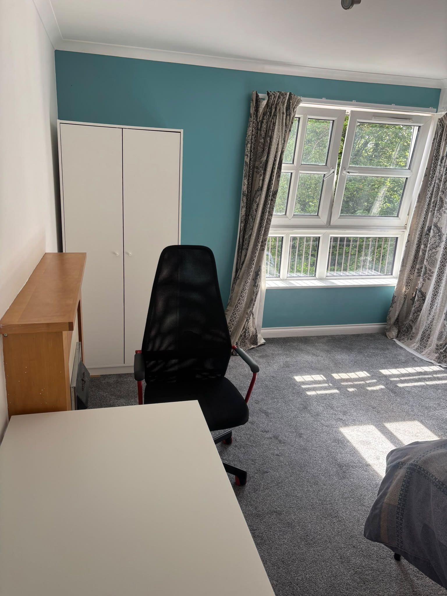 A room with teal walls, a white desk, a black office chair, a wardrobe, and a window with curtains.