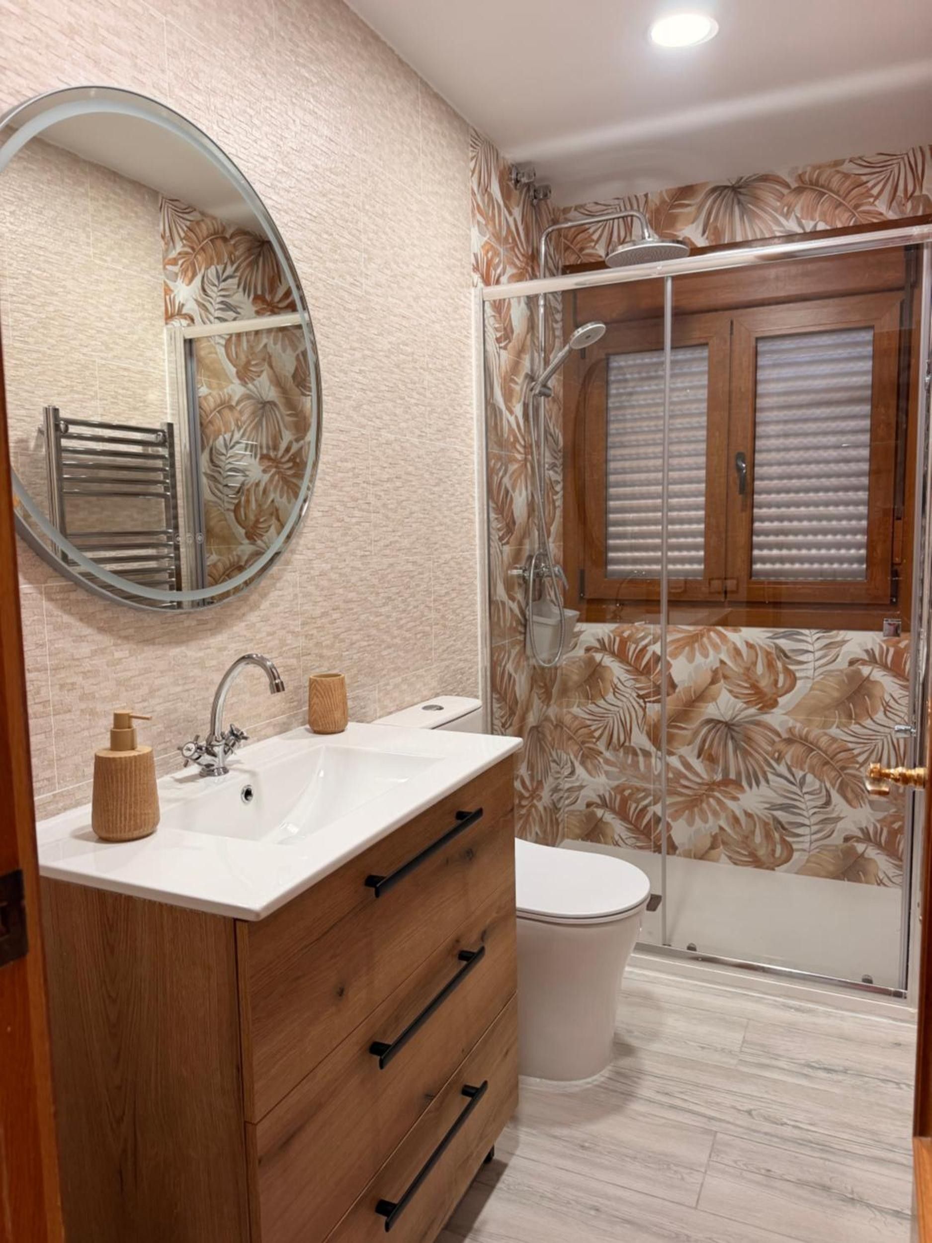 Een badkamer met een houten wastafelmeubel, een ronde spiegel, een douche met glazen deur en een toilet.