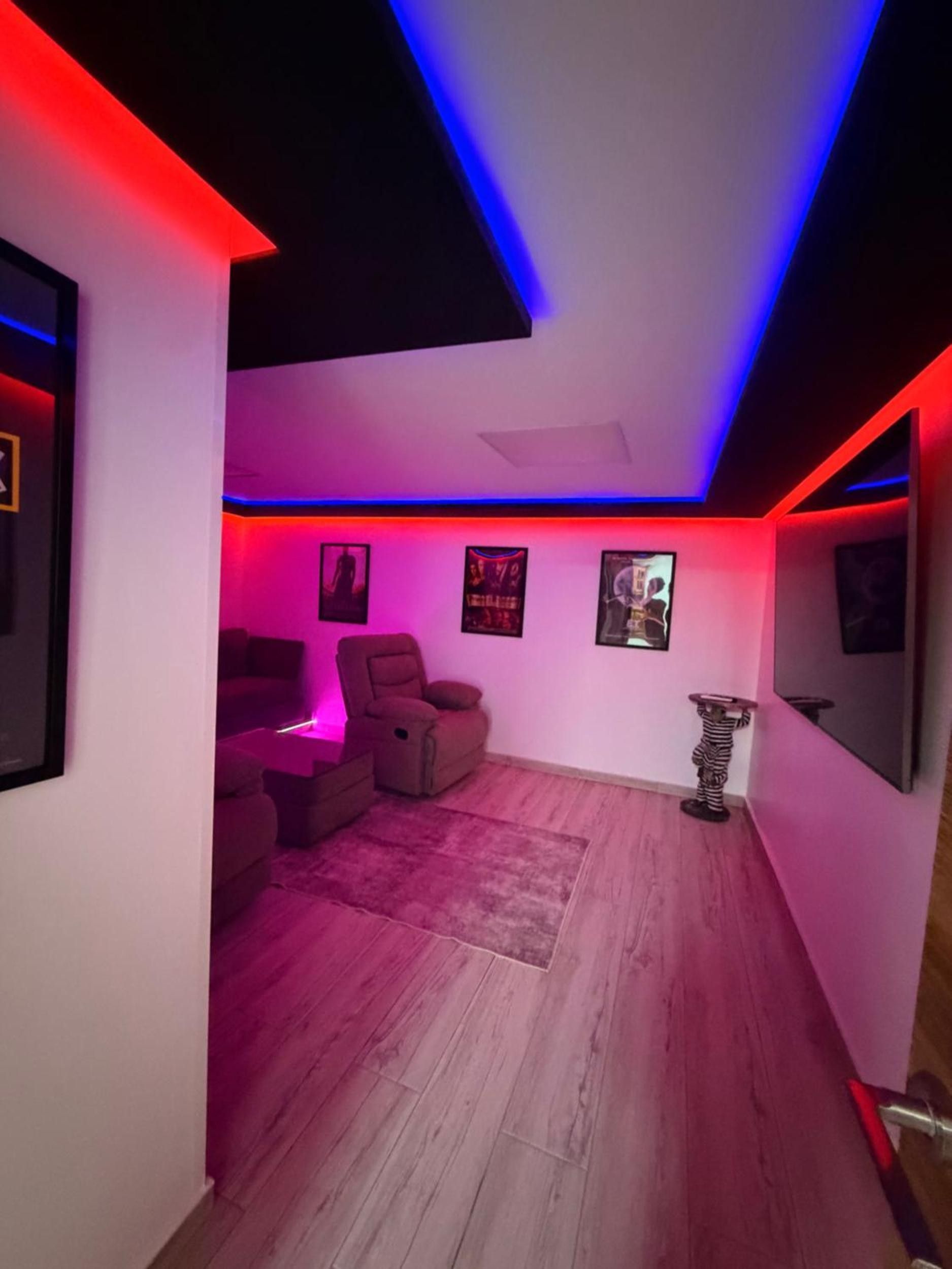 Una stanza con una striscia di luci al neon rossa e blu, una poltrona reclinabile, un tavolino e un televisore a schermo piatto montato a parete.