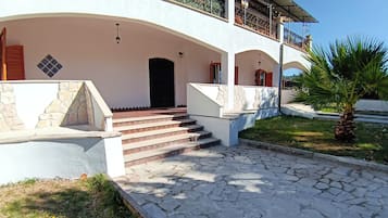 Ein Gebäude mit einer Steintreppe, die zu einer schwarzen Tür führt, einem überdachten Balkon mit Metallgeländern und einer Palme im Vordergrund.