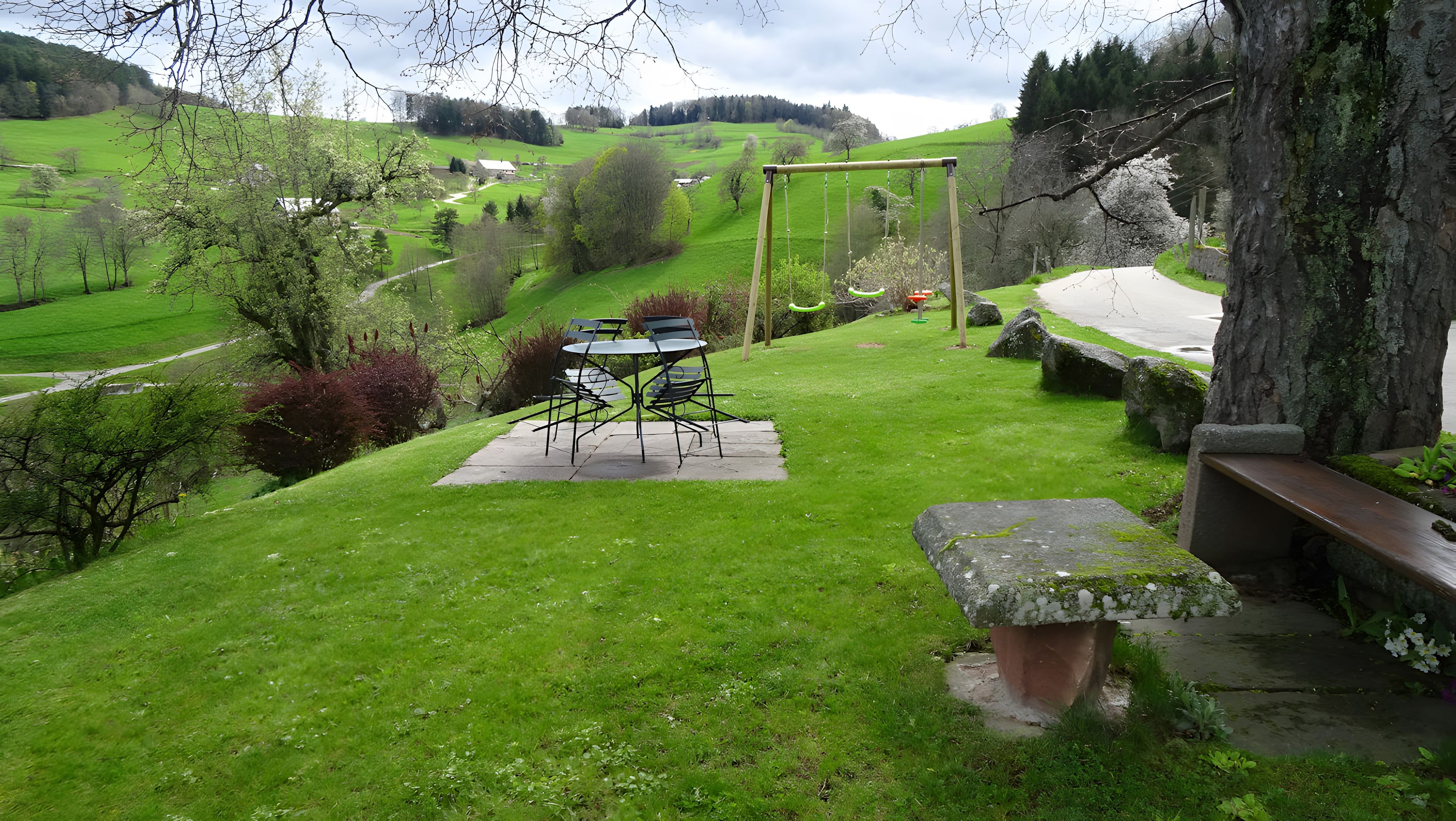 Un espace extérieur verdoyant avec une table et des chaises, un banc en pierre et une vue sur une vallée verdoyante.