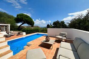 Un área de piscina en la azotea con una piscina azul cristalina, mobiliario exterior blanco y vistas del paisaje circundante.