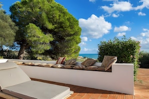 Un espacio exterior con mesas iluminado por el sol, vistas al mar, un gran árbol y una pared blanca.
