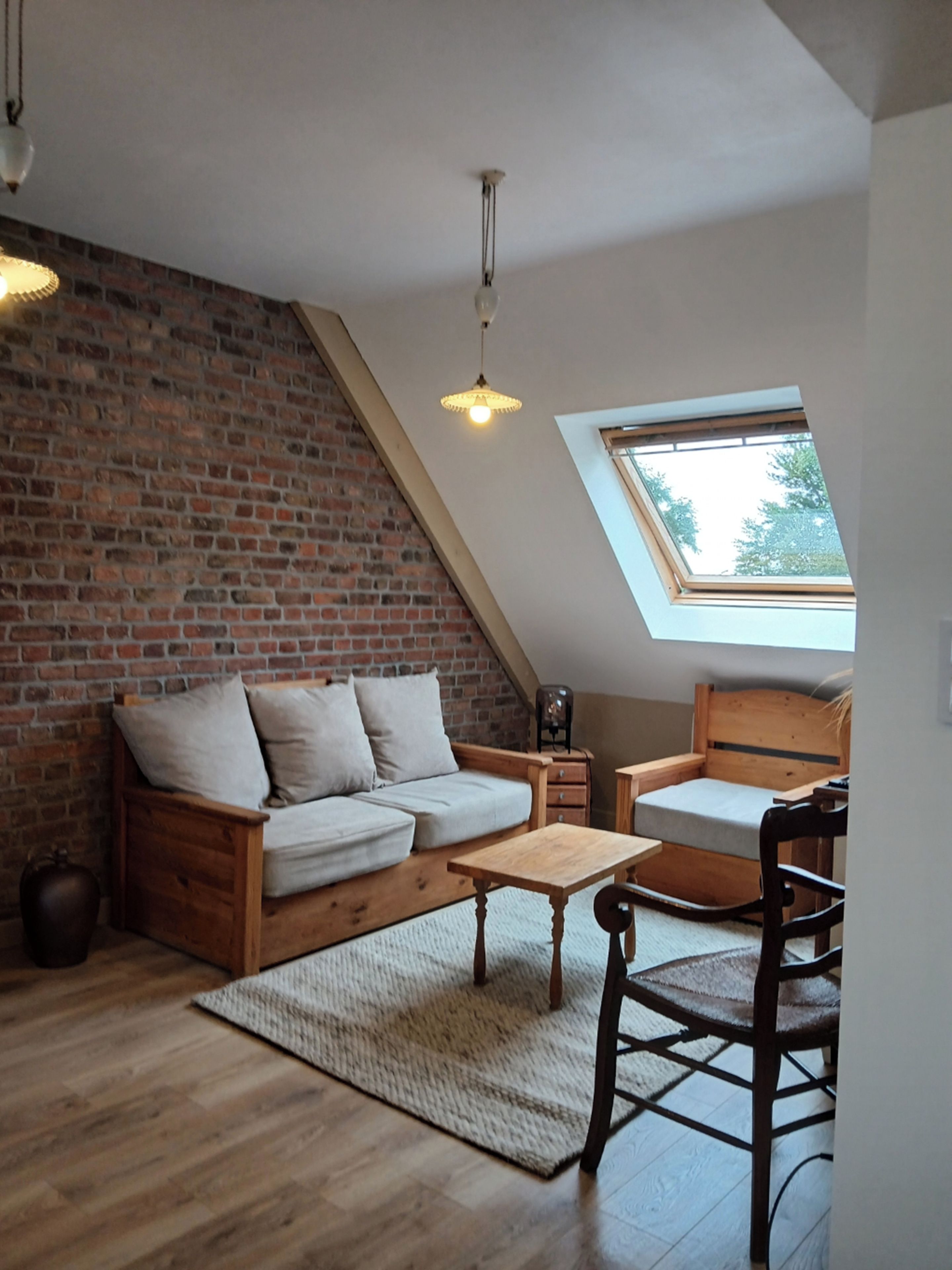 Een loft-achtige kamer met een schuine kap, een openliggende bakstenen muur, houten meubilair en een dakraam.