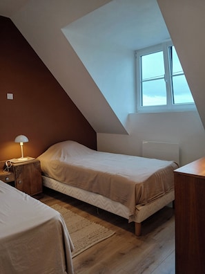 A small, cozy room with a sloped ceiling, a single bed, a bedside table with a lamp, and a window providing natural light.
