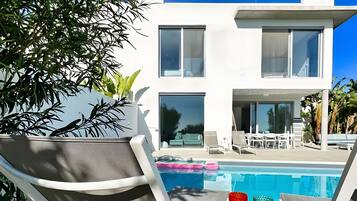 A modern two-story building with a pool, lounge chairs, and a table with glasses.