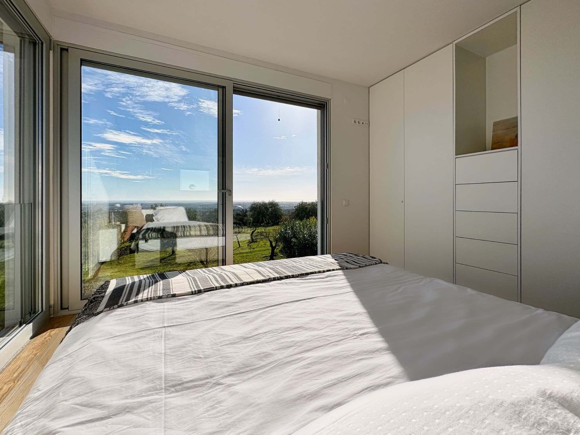 Ein Schlafzimmer mit einer großen Schiebeglastür, die auf einen Balkon führt, einem Bett mit gestreifter Bettdecke und einem weißen Kleiderschrank.