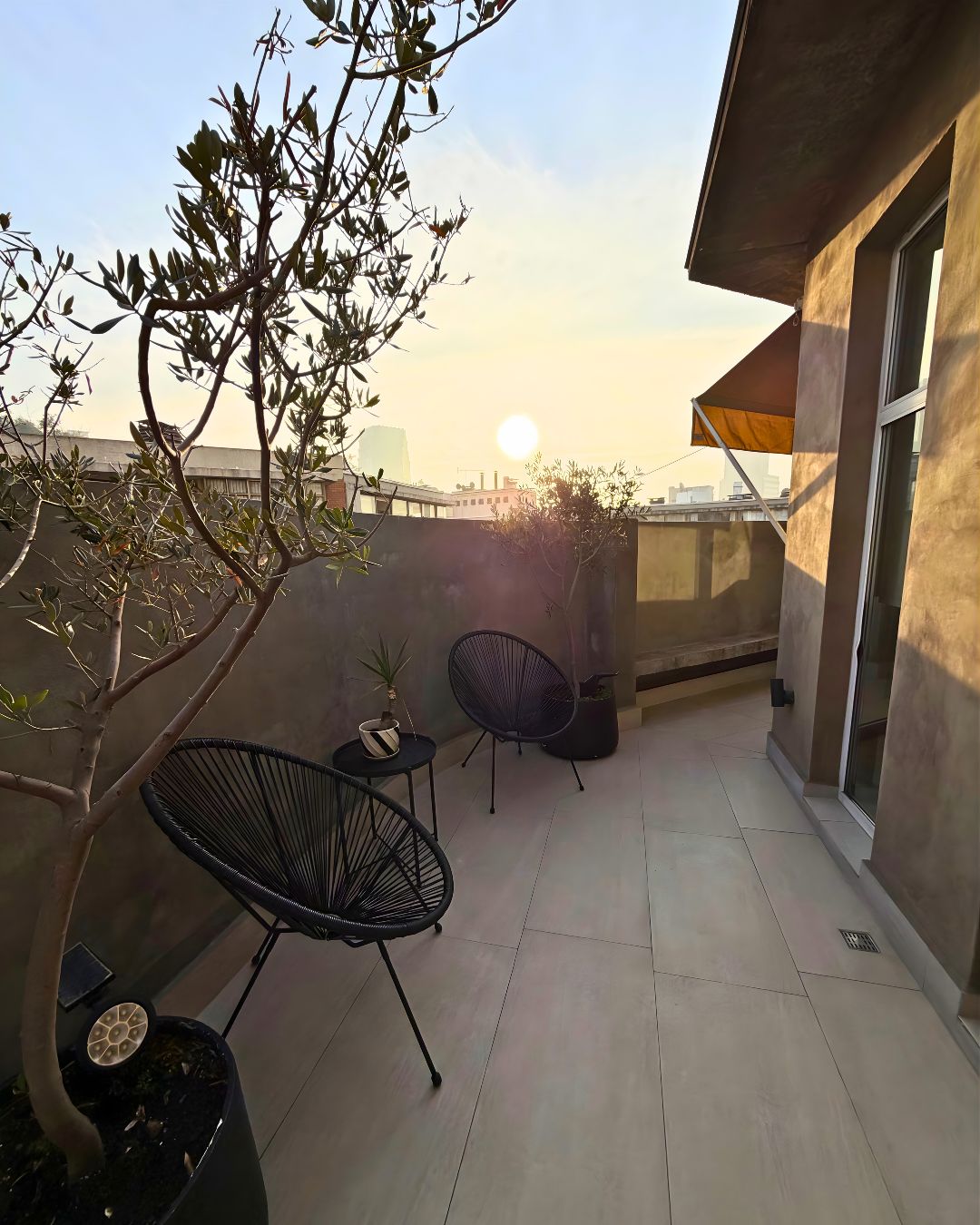 A rooftop terrace with a view of the city skyline, two black chairs, and a potted plant.