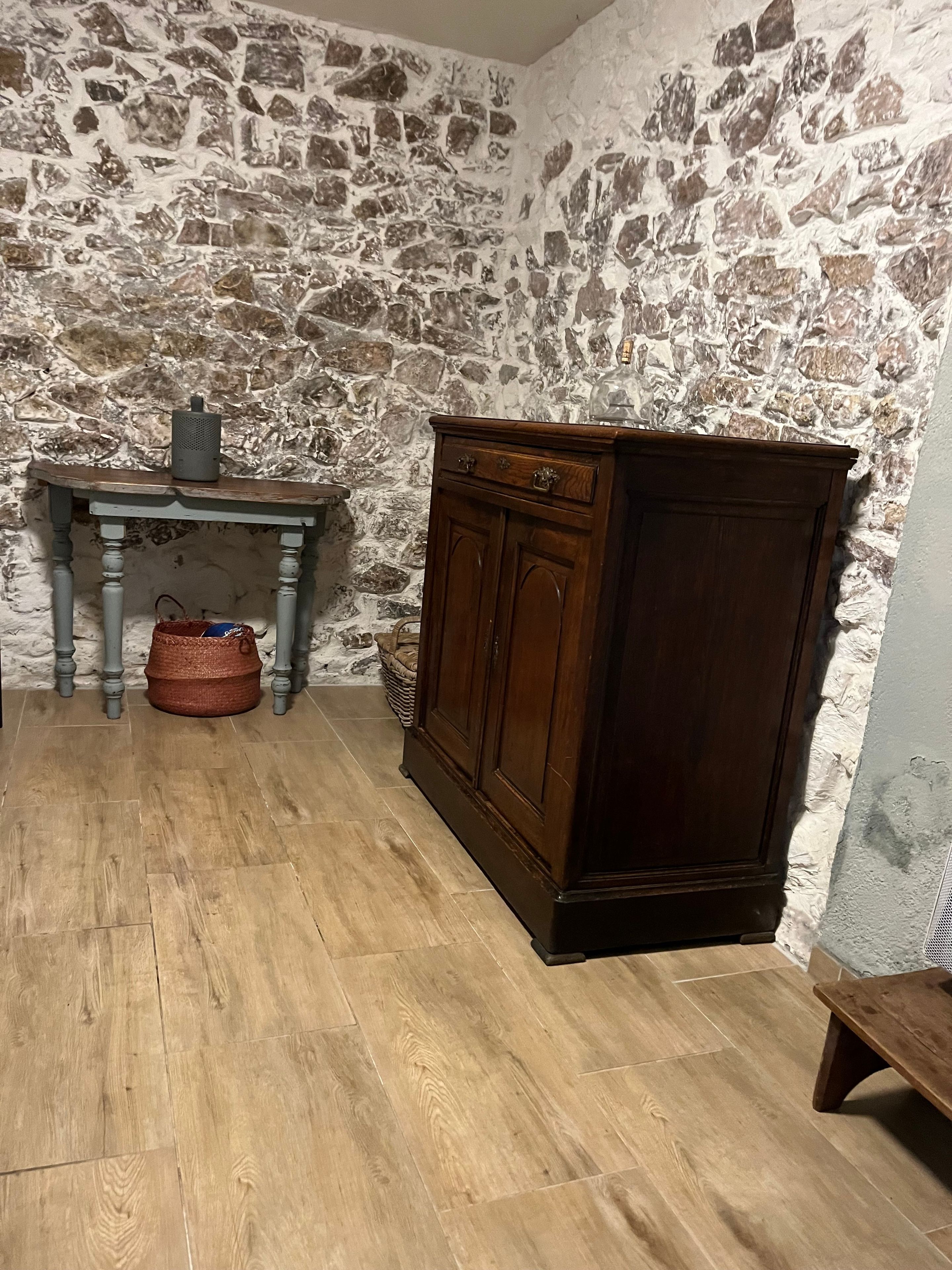 Une pièce avec un mur en pierre, un meuble en bois, une table avec un panier et un banc.