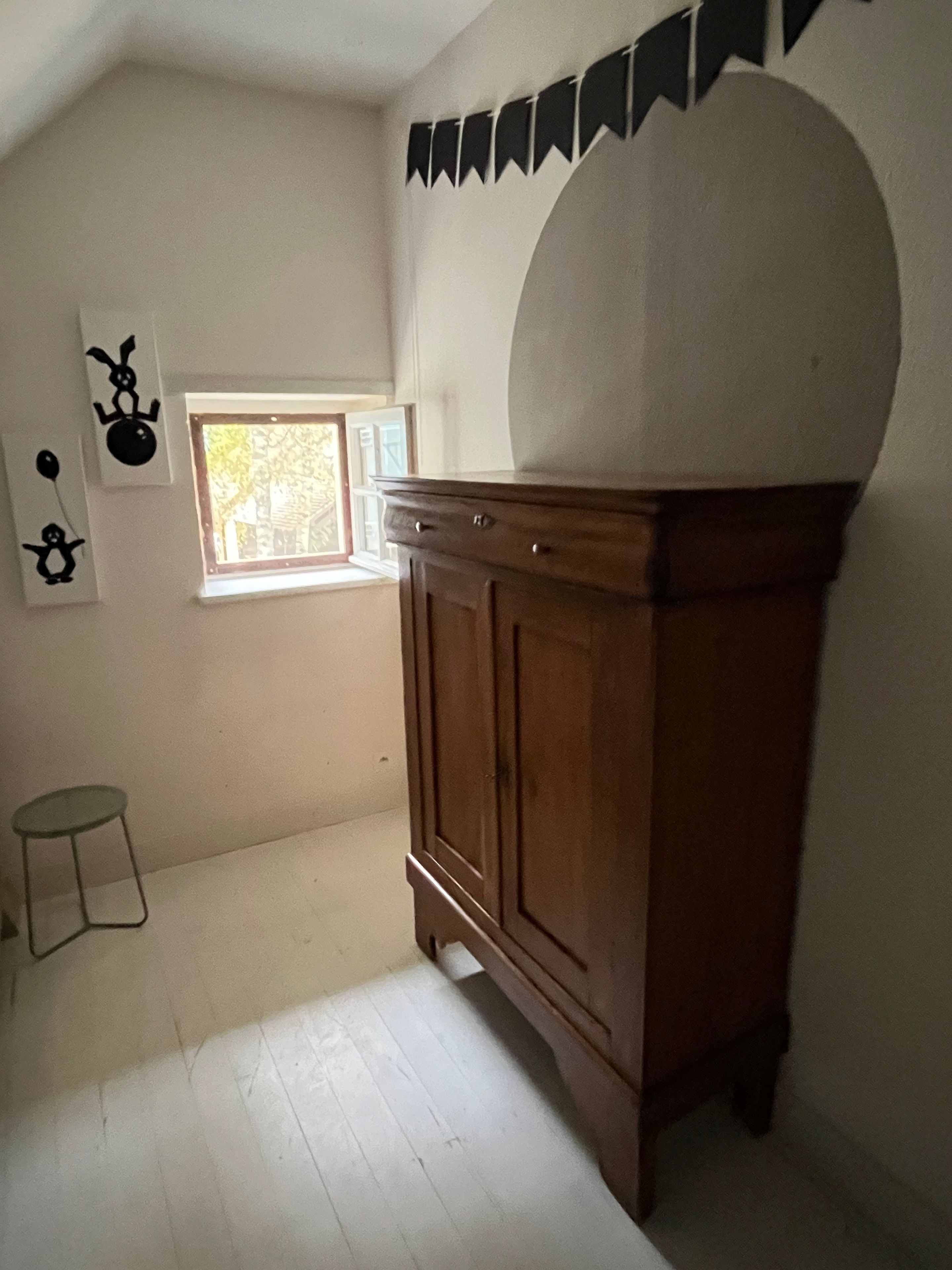 Una habitación con un armario de madera, un taburete pequeño, una ventana con vistas y adornos en la pared.