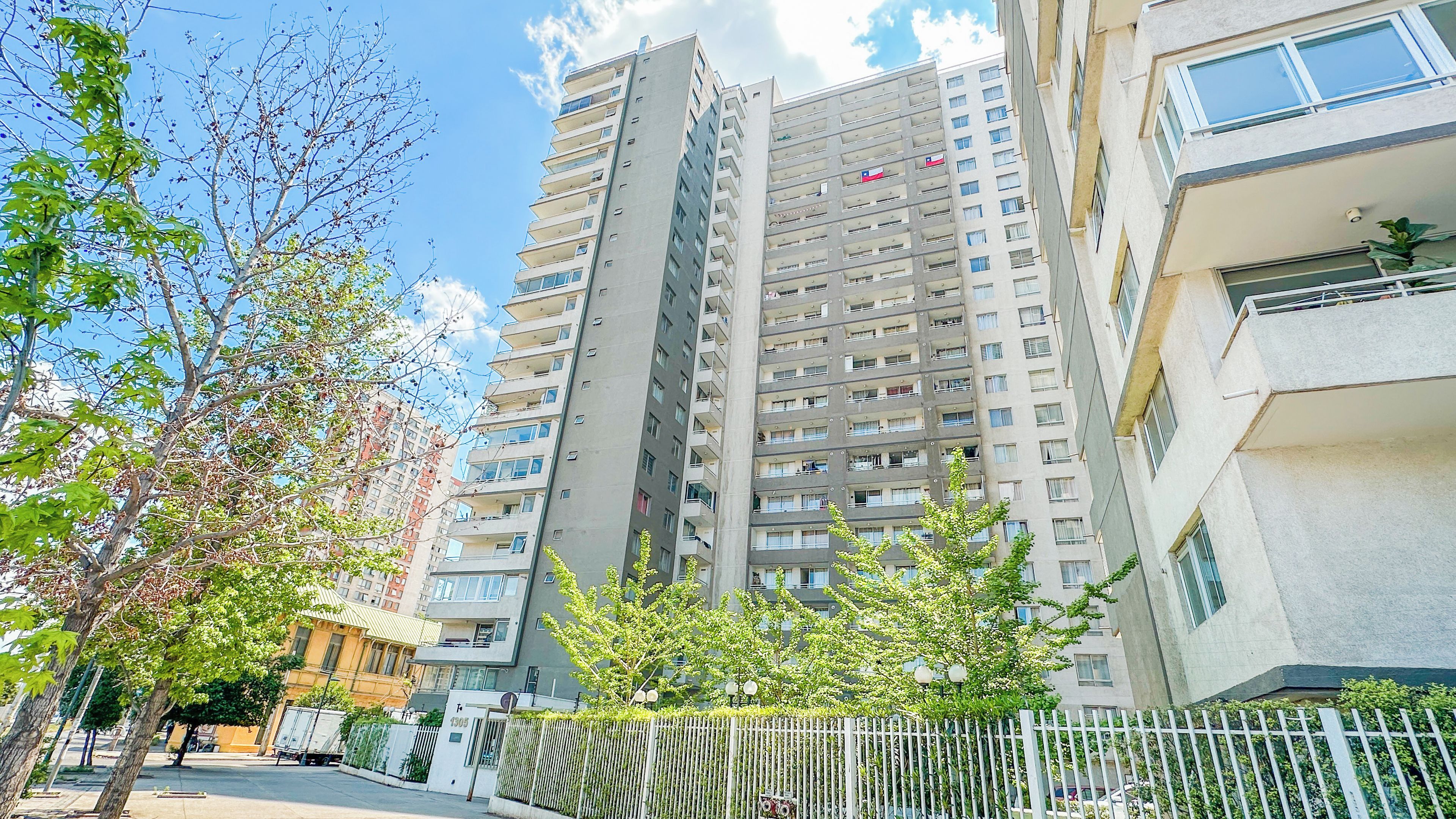 Un edificio residencial alto con varios balcones, entrada protegida y cielo despejado.