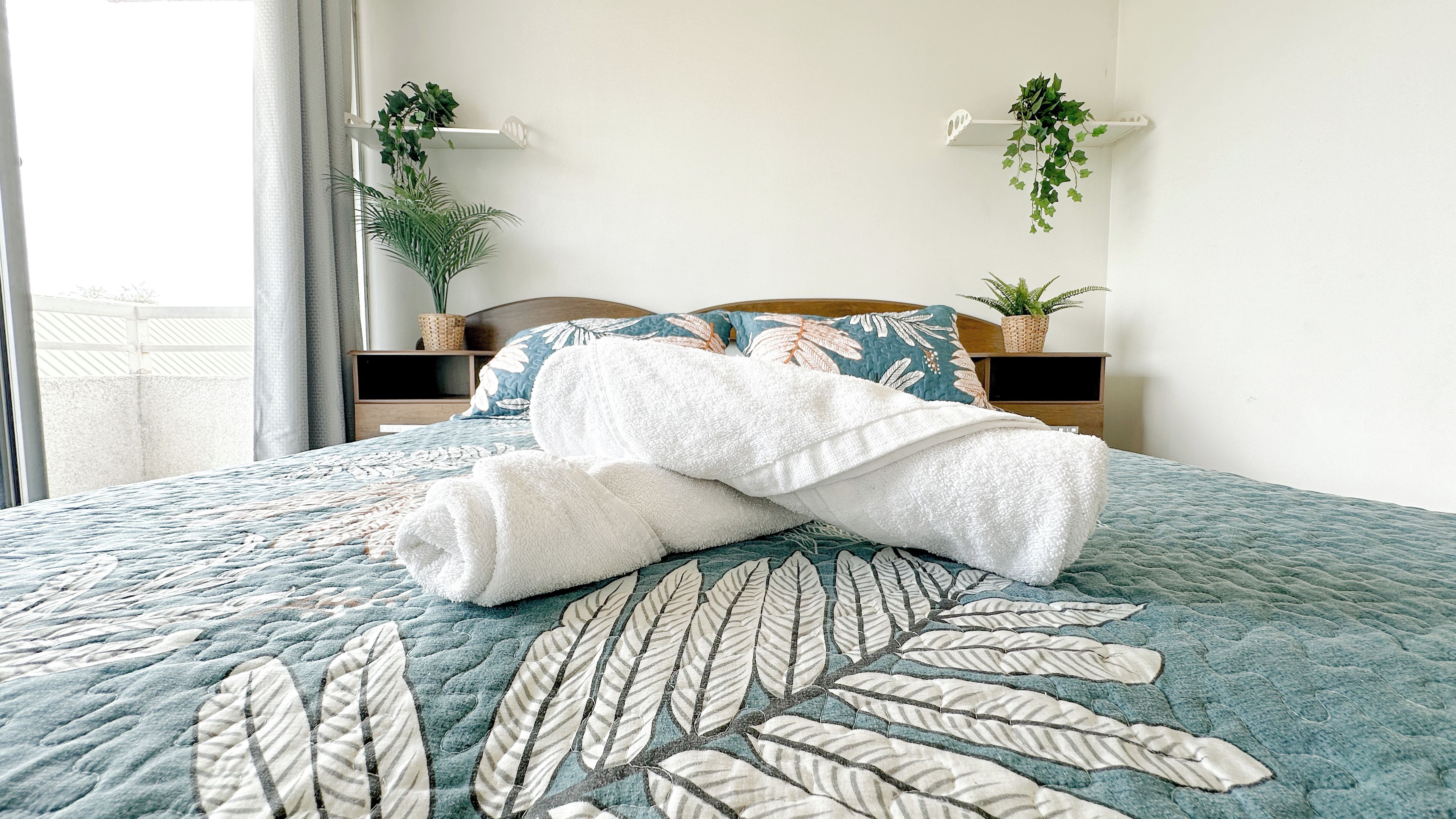 Una cama bien hecha con cubrecama estampado con hojas, un cabecero de madera y dos mesitas de noche con plantas en macetas.